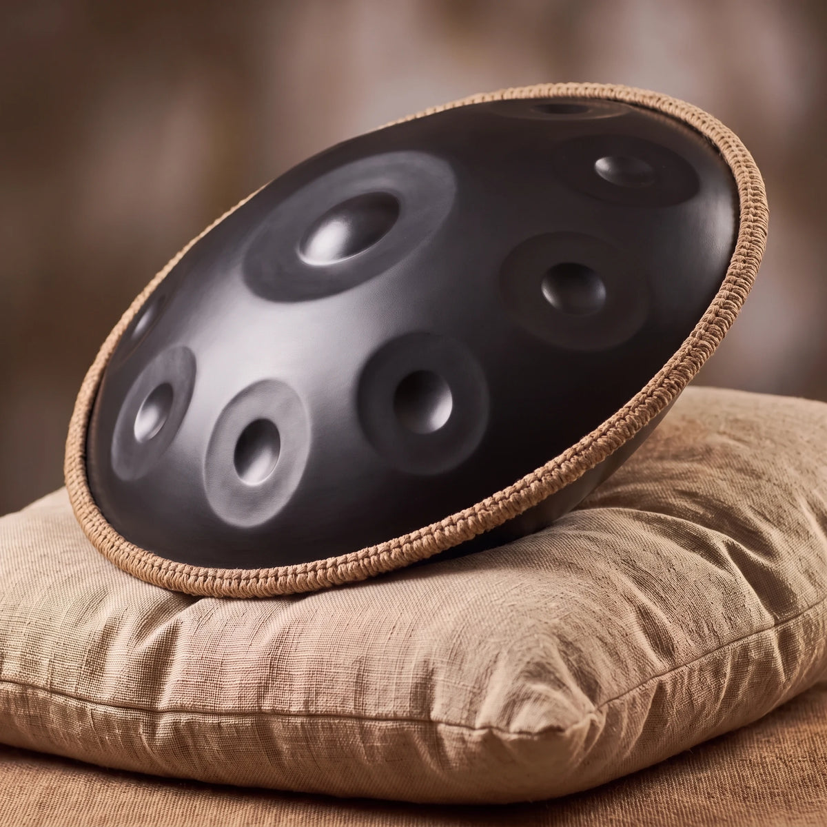 Handpan G minor KURO noir mat avec bord tressé posé de profil sur un coussin en lin – Kovapan