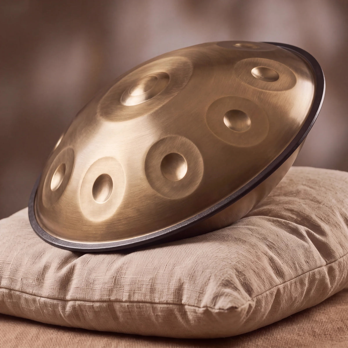 Handpan Sol mineur doré Kovapan bronze satiné – posé de profil sur coussin lin fond bokeh chaud